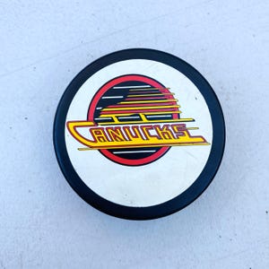 May include: A black and white hockey puck with the Vancouver Canucks logo. The logo features the word "CANUCKS" in yellow, with red and black details. The puck is circular and has a white central area.