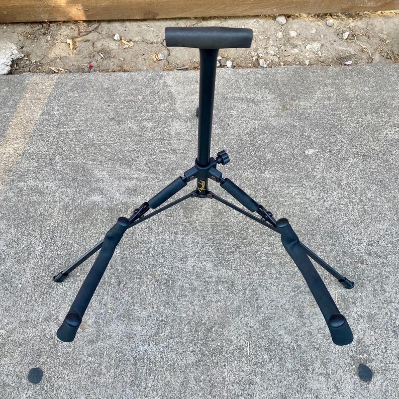 Puede incluir: Un soporte de guitarra de metal negro con base de tr&iacute;pode y brazos de apoyo acolchados con espuma. El poste vertical central cuenta con un reposacabezas en forma de T y un peque&ntilde;o logotipo amarillo de Fender en el eje central.