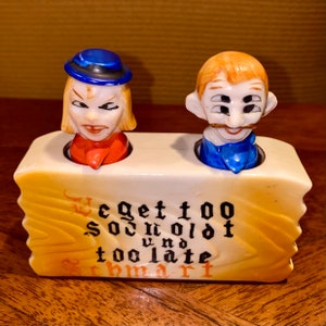 Puede incluir: Un juego de salero y pimentero de cerámica vintage con dos caras de dibujos animados. Una es una mujer con cabello rubio y un sombrero azul, la otra un hombre con cabello naranja y un bigote. La base tiene el texto "We get too old und too late schmart."