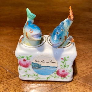 Puede incluir: Juego de salero y pimentero vintage de cerámica. Presenta dos peces azules con detalles naranjas y verdes. La base blanca tiene un diseño floral y una imagen circular con el texto "New Haven, Conn."
