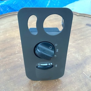 Puede incluir: Un panel de interruptor de faros de automóvil negro con un dial giratorio y un control deslizante. El panel tiene dos recortes circulares e iconos para faros y luces antiniebla. El panel está hecho de plástico.