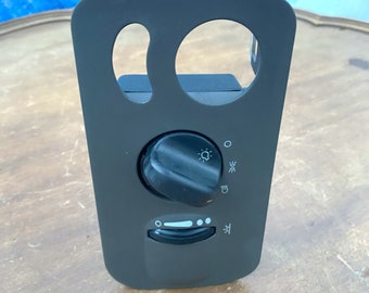 May include: A black automotive headlight switch panel with a rotary dial and a slider. The panel has two circular cutouts and icons for headlights and fog lights. The panel is made of plastic.