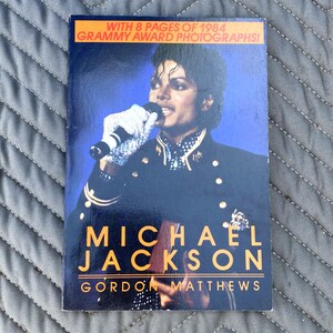 May include: A book cover featuring Michael Jackson in a black jacket with gold accents, holding a microphone. The title "MICHAEL JACKSON" is in large orange letters, with "GORDON MATTHEWS" below. The cover includes the text "WITH 8 PAGES OF 1984 GRAMMY AWARD PHOTOGRAPHS!"