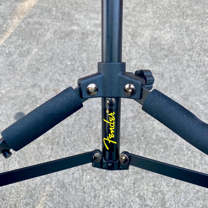 Puede incluir: Un soporte de guitarra de metal negro con un poste vertical central que tiene el logotipo de Fender en amarillo impreso. El soporte incluye dos brazos de apoyo cubiertos con espuma negra, conectados por un eje central con remaches plateados.