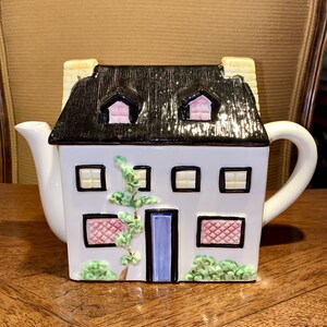 以下が含まれることがあります： 黒い屋根、黄色の煙突、青いドアが特徴の、家型の白いセラミックティーポット。家にはピンクと黄色のアクセントが付いた窓があり、緑のつるが側面を這っています。ハンドルと注ぎ口は白です。