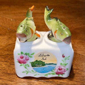 Puede incluir: Un juego de salero y pimentero de cerámica vintage con dos saleros en forma de pez en una base blanca. Los peces son verdes con detalles en naranja y negro. La base tiene un diseño floral y una pequeña imagen con el texto "Lafayette Ind."