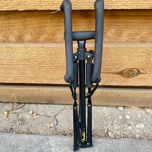Puede incluir: Un soporte de guitarra plegable negro con brazos de apoyo acolchados con espuma y un poste vertical central. El soporte presenta un logotipo Fender amarillo impreso cerca de la base, con bisagras y remaches met&aacute;licos que unen los componentes.