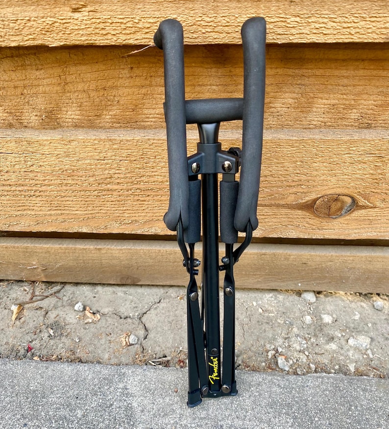 Puede incluir: Un soporte de guitarra plegable negro con brazos de apoyo acolchados con espuma y un poste vertical central. El soporte presenta un logotipo Fender amarillo impreso cerca de la base, con bisagras y remaches met&aacute;licos que unen los componentes.