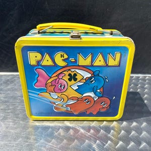 May include: Vintage Pac-Man lunchbox with a yellow handle and trim. The front features the Pac-Man logo in yellow letters on a blue background, along with images of Pac-Man and ghosts. The lunchbox has a metal clasp.