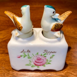 Puede incluir: Un juego de salero y pimentero de cerámica vintage. El juego presenta dos saleros con forma de pájaro sobre una base blanca. La base está decorada con una rosa rosa y las palabras "Salt" y "Pepper". Los pájaros tienen detalles azules, marrones y blancos.