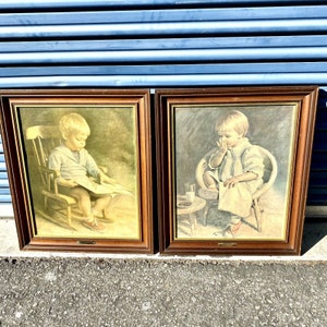 May include: Two framed vintage prints. One shows a young child in a rocking chair reading a book. The other shows a child in a wicker chair. Both prints have a brown wooden frame and are displayed on a concrete surface.