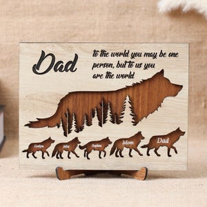 May include: Wooden plaque with a wolf design and the word "Dad" in a script font. The plaque includes the quote "to the world you may be one person, but to us you are the world." Below the wolf are smaller wolf figures with names.