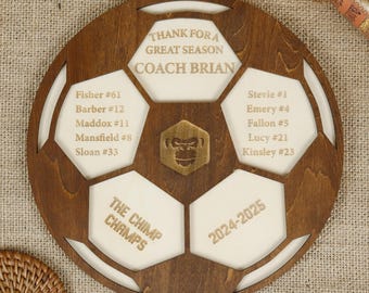 May include: A wooden soccer ball plaque with a white background and gold lettering. The plaque features the text "Thank for a Great Season Coach Brian", "The Chimp Champs", and "2024-2025". The plaque also includes a list of names and numbers.