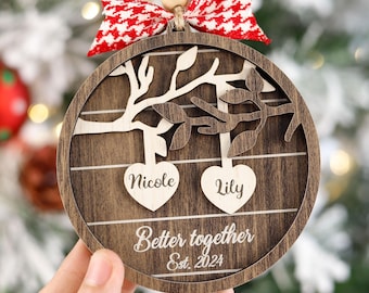 May include: A round wooden Christmas ornament with a tree design and two hearts with the names "Nicole" and "Lily" on them. The ornament also says "Better together Est. 2024".