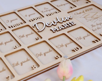 May include: A wooden photo frame with slots for pictures from ages 3 to 11th grade. The frame is personalized with the name "Devlin Marie" and the text "Class of 2039".