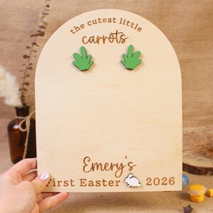 May include: A wooden Easter-themed sign with the text "the cutest little carrots" and two green carrot tops. The sign also includes the name "Emery's", "First Easter", a bunny, and the year 2026.