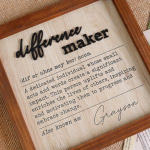 May include: A wooden sign with the word "difference maker" in black lettering. The definition of the word is written below, followed by the name "Grayson".