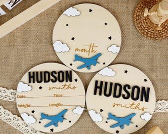 Puede incluir: Tres discos de madera para hitos de bebé con un avión azul y un diseño de nubes blancas. El disco superior dice "mes" en letras doradas. Los dos discos inferiores dicen "HUDSON smiths" en letras negras y tienen espacios para escribir la fecha, la hora, el peso y la longitud.