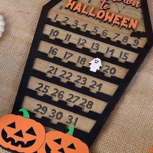 May include: A wooden countdown calendar shaped like a coffin with the text "Countdown to Halloween" and numbers 1 through 31. The calendar is decorated with a spider web, a spider, and a ghost.