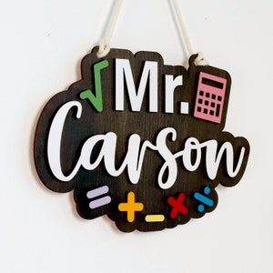 May include: A dark brown wooden sign with the text "Mr. Carson" in white lettering. The sign has a green square root symbol, a pink calculator, and colorful math symbols.
