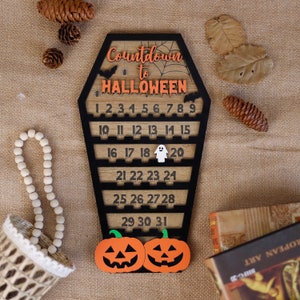 May include: Wooden Halloween countdown calendar shaped like a coffin with a black and orange color scheme. The calendar features a spider web design and the numbers 1 through 31. The calendar is decorated with two orange pumpkins with black eyes and a white ghost.