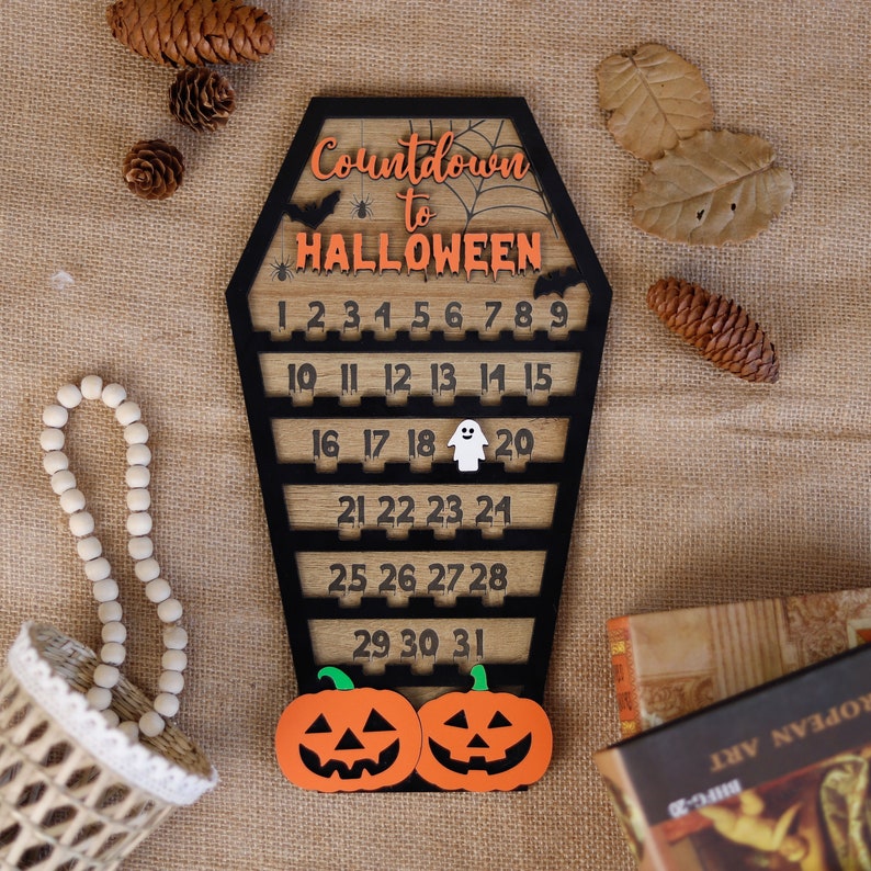 May include: Wooden Halloween countdown calendar shaped like a coffin with a black and orange color scheme. The calendar features a spider web design and the numbers 1 through 31. The calendar is decorated with two orange pumpkins with black eyes and a white ghost.