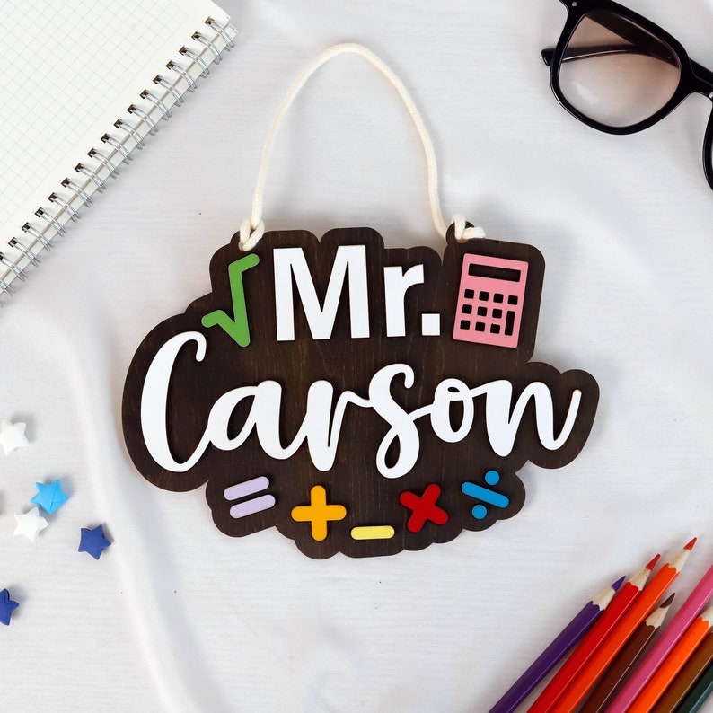 May include: A dark brown wooden sign with the text "Mr. Carson" in white lettering. The sign has a green checkmark, a pink calculator, and colorful math symbols.