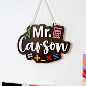 May include: A wooden sign with the text "Mr. Carson" in white lettering. The sign is decorated with a calculator, a square root symbol, and mathematical symbols including an equals sign, plus sign, minus sign, multiplication sign, and division sign.