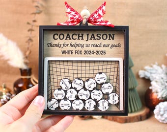 May include: A personalized soccer coach gift featuring a wooden frame with a soccer net and miniature soccer balls. The frame includes the text "COACH JASON" and "WHITE FOX 2024-2025". Each ball has a player's name and number. A red and white bow adorns the top.