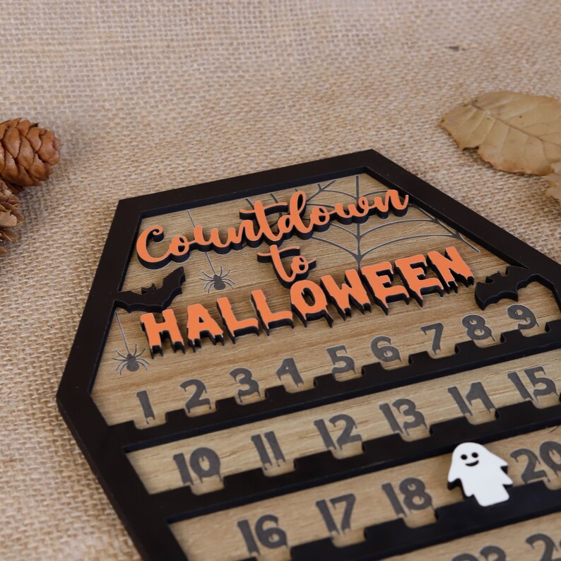 May include: A wooden countdown calendar for Halloween. The calendar is shaped like a coffin and has a black frame. The numbers 1 through 20 are displayed on the calendar, with a ghost figure at the bottom. The text "Countdown to Halloween" is written in orange letters on the top of the calendar.