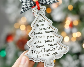 May include: A wooden Christmas tree ornament with a silver glitter outline. The ornament has names engraved on it, including "Emily," "Brian," and "Jessica." It is topped with a black and white bow and the text "Mrs. Christopher Pre-K 2023-2024."