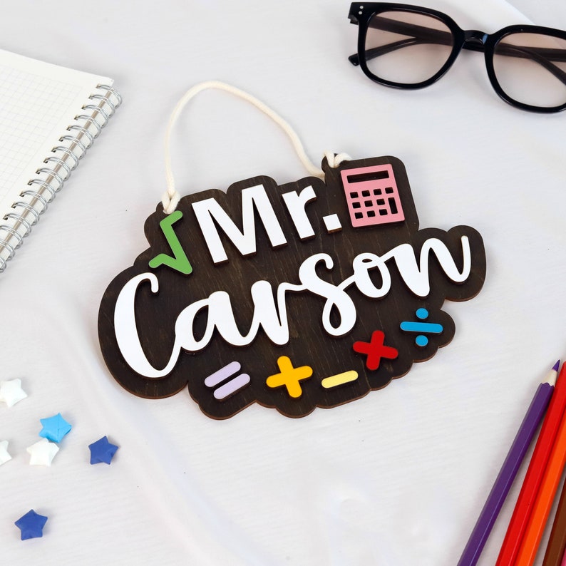 May include: A brown wooden sign with the text "Mr. Carson" in white lettering. The sign has a pink calculator, a green check mark, and colorful math symbols.