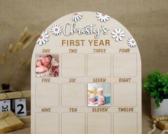 May include: A wooden photo display with a scalloped top decorated with white daisies. The display has 12 spaces for photos, labeled with the numbers one through twelve. The text "Christy's First Year" is written in gold lettering on the top of the display.