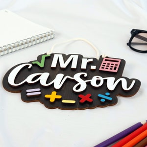 May include: A wooden sign with the text "Mr. Carson" in white lettering. The sign is decorated with a green checkmark, a pink calculator, and colorful math symbols.