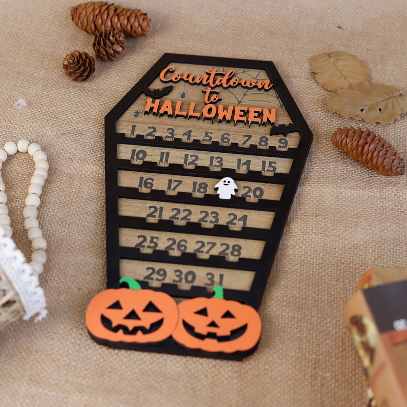 May include: A wooden countdown calendar shaped like a coffin with the text "Countdown to HALLOWEEN" and numbers 1 through 31. The calendar has a black background and orange numbers. There are two jack-o'-lantern pumpkins at the bottom of the calendar.