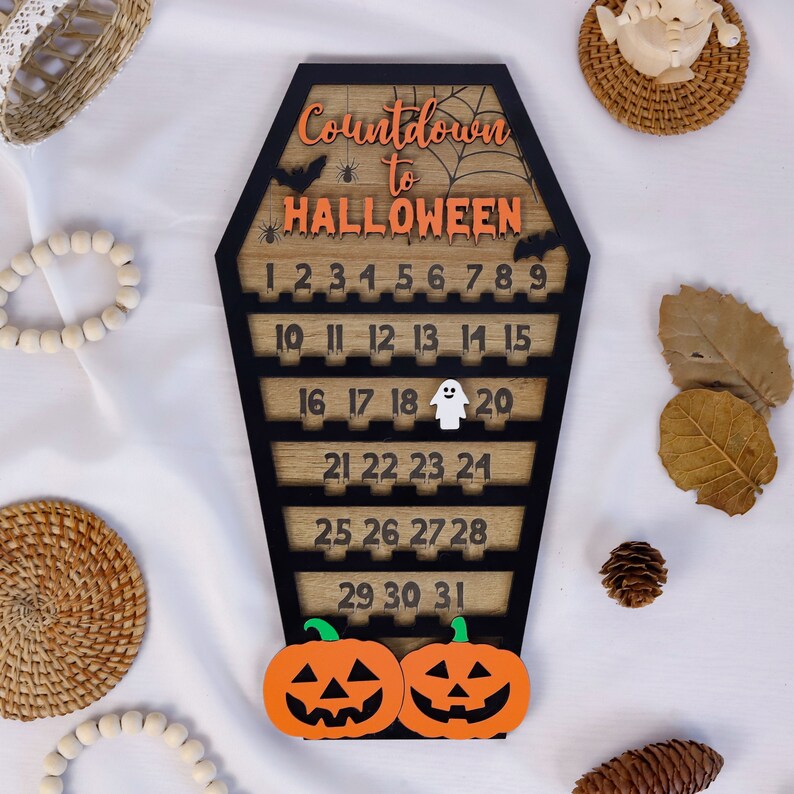 May include: A wooden countdown calendar shaped like a coffin with the text "Countdown to HALLOWEEN" in orange and black lettering. The calendar has numbers 1 through 31 on it, with a ghost on the 18th and two pumpkins at the bottom.