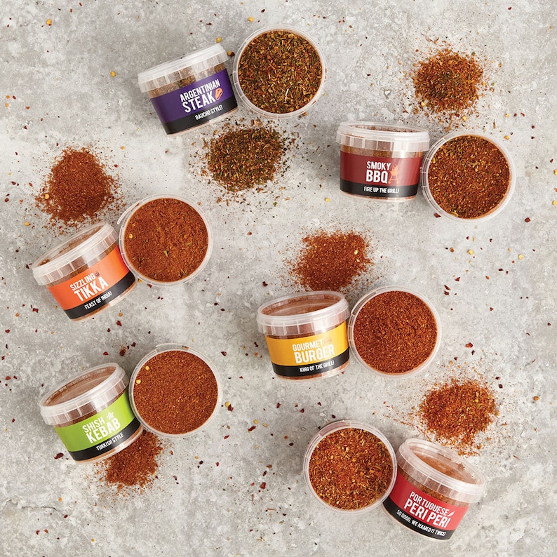 May include: Ten small glass jars filled with different spice blends. The jars are arranged in a circle on a gray background. The labels on the jars include the names of the spice blends, such as "Sizzling Tikka", "Gourmet Burger", and "Smoky BBQ".