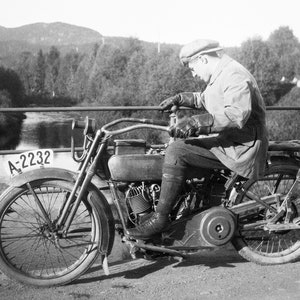 Biker on road trip riding his Harley Davidson motorcycle in the early 1920's. Picture is taken on a bridge crossing a river. Restored vintage black and white picture available as museum quality giclee print.