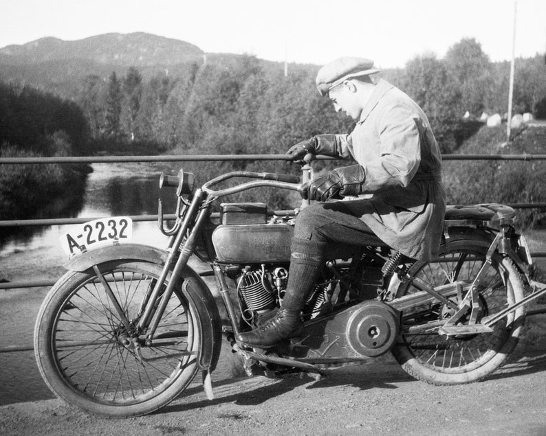 Biker on road trip riding his Harley Davidson motorcycle in the early 1920's. Picture is taken on a bridge crossing a river. Restored vintage black and white picture available as museum quality giclee print.