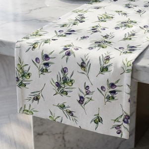 May include: White table runner with a repeating pattern of olive branches with green and purple olives. The runner is placed on a marble table. A small, round, white dish with gold-colored objects sits on the table.