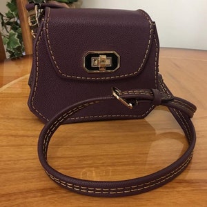 May include: A small, purple leather crossbody bag with a gold clasp and a long strap. The bag has a stitched design around the edges.