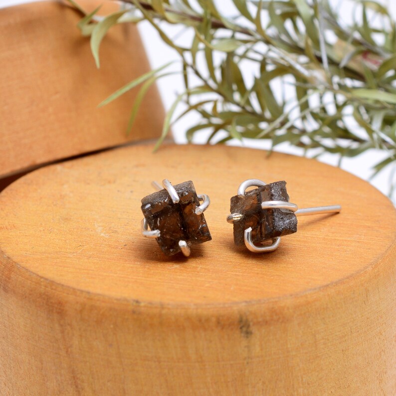 May include: A pair of raw smoky quartz stud earrings. Each earring features a dark brown, cube-shaped quartz crystal wrapped in a silver wire setting. The earrings are displayed on a wooden surface.