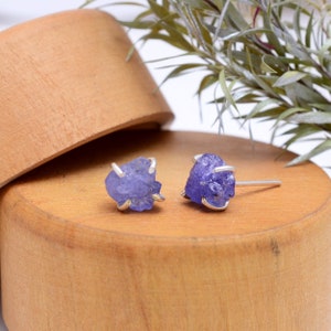 Può includere: Un paio di orecchini a perno in argento con accenti di pietre preziose grezze blu-viola. Gli orecchini sono esposti su una superficie in legno.