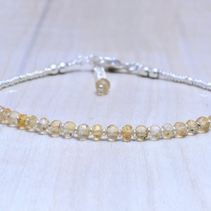 Könnte beinhalten: Ein zartes Armband mit kleinen, facettierten, hellgelben Perlen. Die Perlen sind auf einer silbernen Kette aufgefädelt, mit einem passenden Verschluss. Das Armband wird auf einer hellen Holzoberfläche präsentiert.