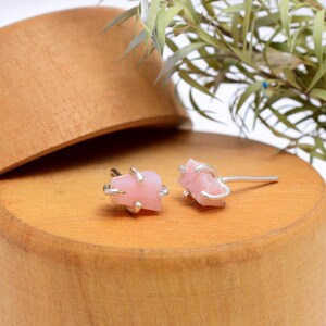 May include: A pair of pink gemstone stud earrings with silver-colored metal accents. The earrings are set on a wooden surface with greenery in the background. The gemstones have a rough, natural appearance.