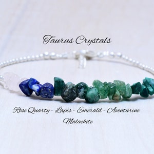 May include: A silver bracelet with a row of gemstone beads. The beads are pink, blue, green, and dark green. The bracelet is labeled "Taurus Crystals" and "Rose Quartz - Lapis - Emerald - Aventurine Malachite".