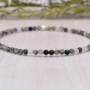 Könnte beinhalten: Ein zartes Armband aus kleinen, runden Perlen in Grautönen, Schwarz und Weiß. Die Perlen sind in einem sich wiederholenden Muster angeordnet, was einen dezenten und stilvollen Look erzeugt.