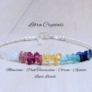 Può includere: Un braccialetto d'argento con un arcobaleno di pietre preziose, tra cui pietra di luna, turmalina rosa, citrino, apatite e lapislazzuli. Il braccialetto è su una superficie di legno bianco. Libra Crystals.
