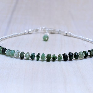 May include: A delicate bracelet featuring a strand of small, faceted emerald beads in varying shades of green. The beads are strung on a silver chain, with a small emerald charm dangling near the clasp. The bracelet is designed for a minimalist aesthetic.
