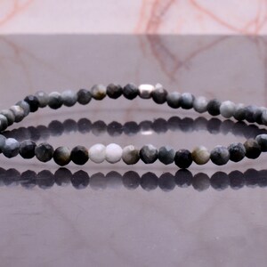 Könnte beinhalten: Ein Perlenarmband mit facettierten, runden Perlen in Grau-, Schwarz- und Weißtönen. Das Armband ist auf einem flexiblen Band aufgezogen und hat einen silberfarbenen Verschluss. Die Perlen haben einen Durchmesser von ca. 0,5 cm.